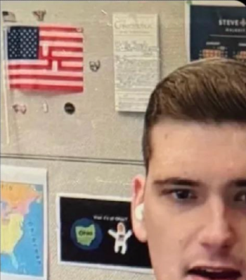 A congressional aide to Congressman Dave Taylor standing in front of a bulletin board. On the board is an American flag in which three of the red stripes have been altered to form a swastika in the middle of the flag