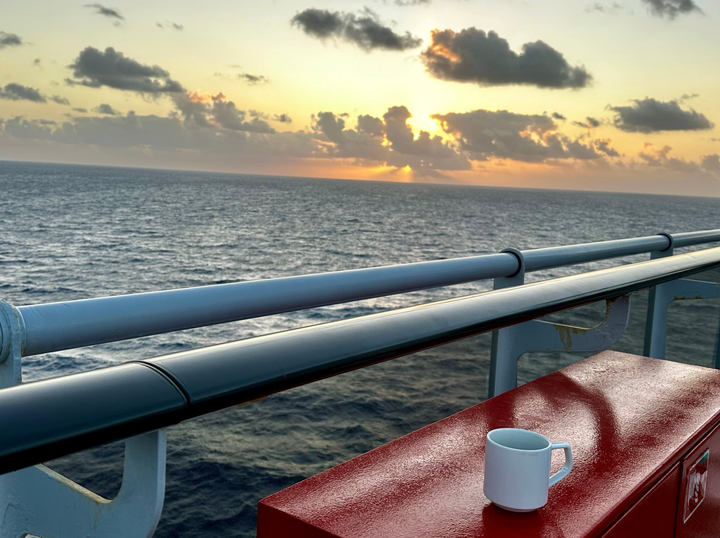 A cup of coffee on a ship's ledge with the ocean and the sunrise in the background.